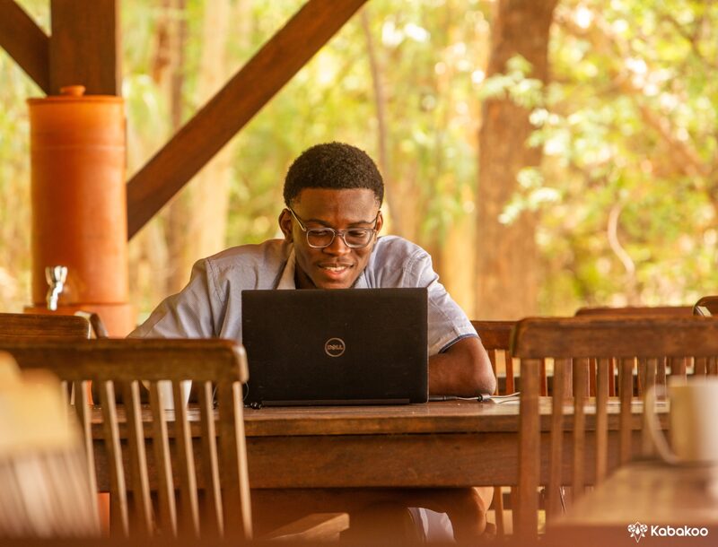 A learner works on a laptop at a wooden table on a shaded terrace, a clay water pot beside him.
