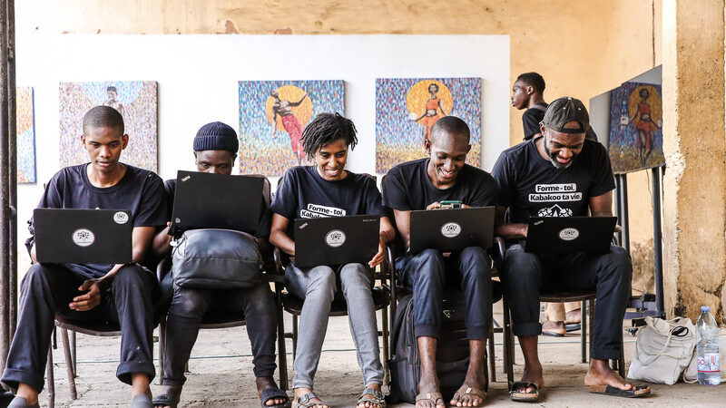 Five team members work on laptops outside at La Maison de l'Étonnement, colorful paintings on the wall behind them.