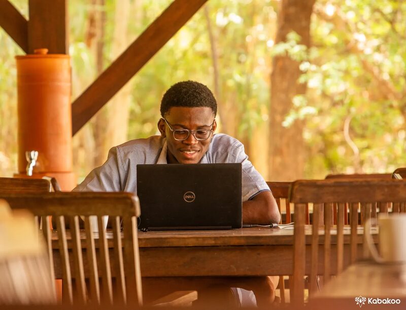A Kabakoo learner works on a laptop at a wooden table outdoors, next to a terracotta water pot.