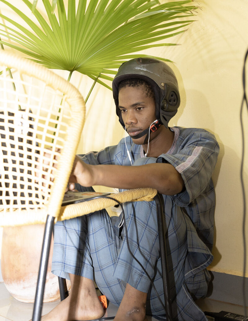 Seydou Ba sits barefoot on the floor wearing a moto helmet and earphones, debugging code on a laptop balanced on a woven chair.